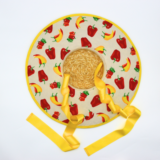 A wide-brimmed straw hat with a chili and pepper fabric design, featuring yellow satin ribbons and metal eyelets.