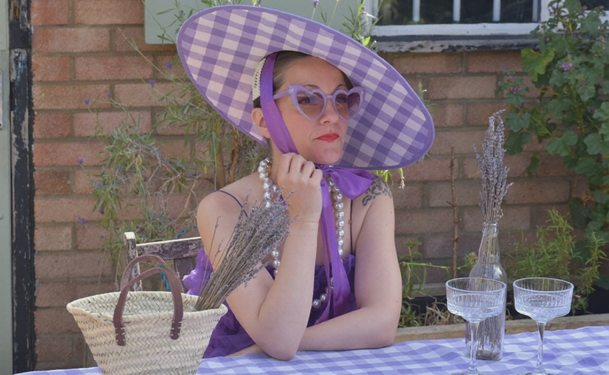Load video: A lady wearing a straw summer hat with gingham lilac fabric, tied up under her chin. She is enjoying a drink at a garden surrounded by flowers and plants